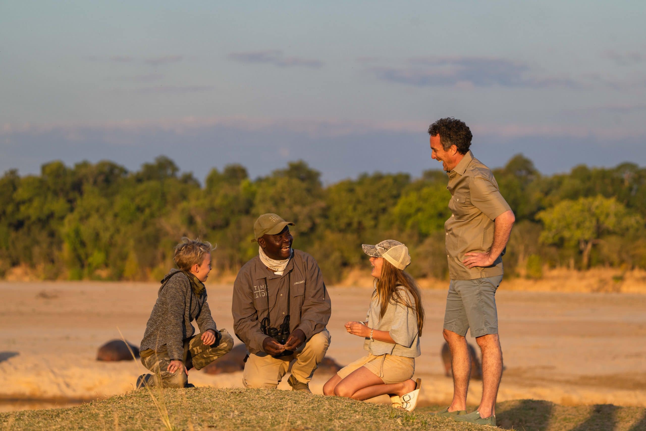 Two children kneeling on the ground with their safari guide and their dad is standing next to them on safari with Time + Tide in Zambia