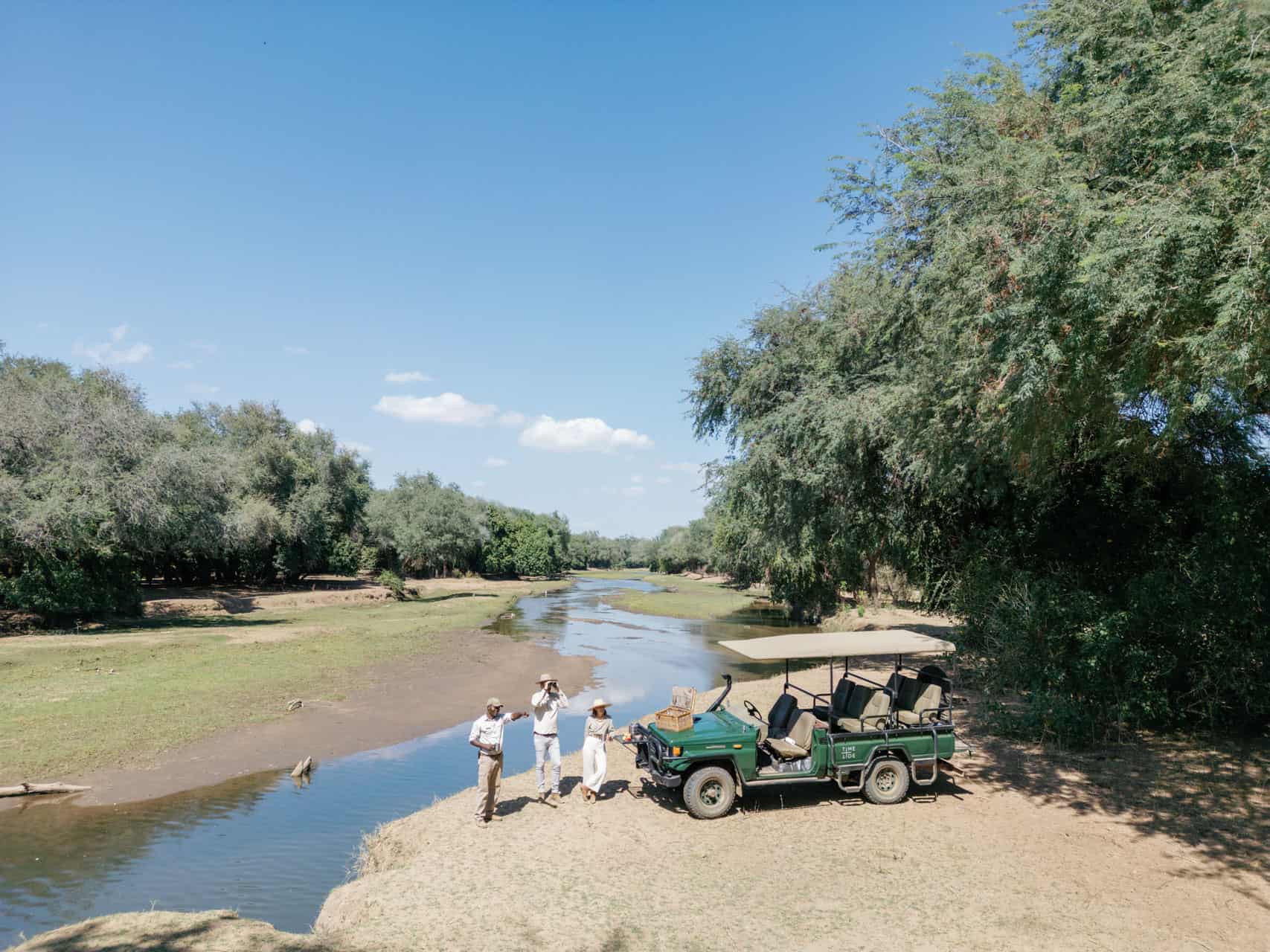 A couple at a morning coffee stop out of the game drive vehicle with their guide along the banks of the river in Zambia with Time + Tide