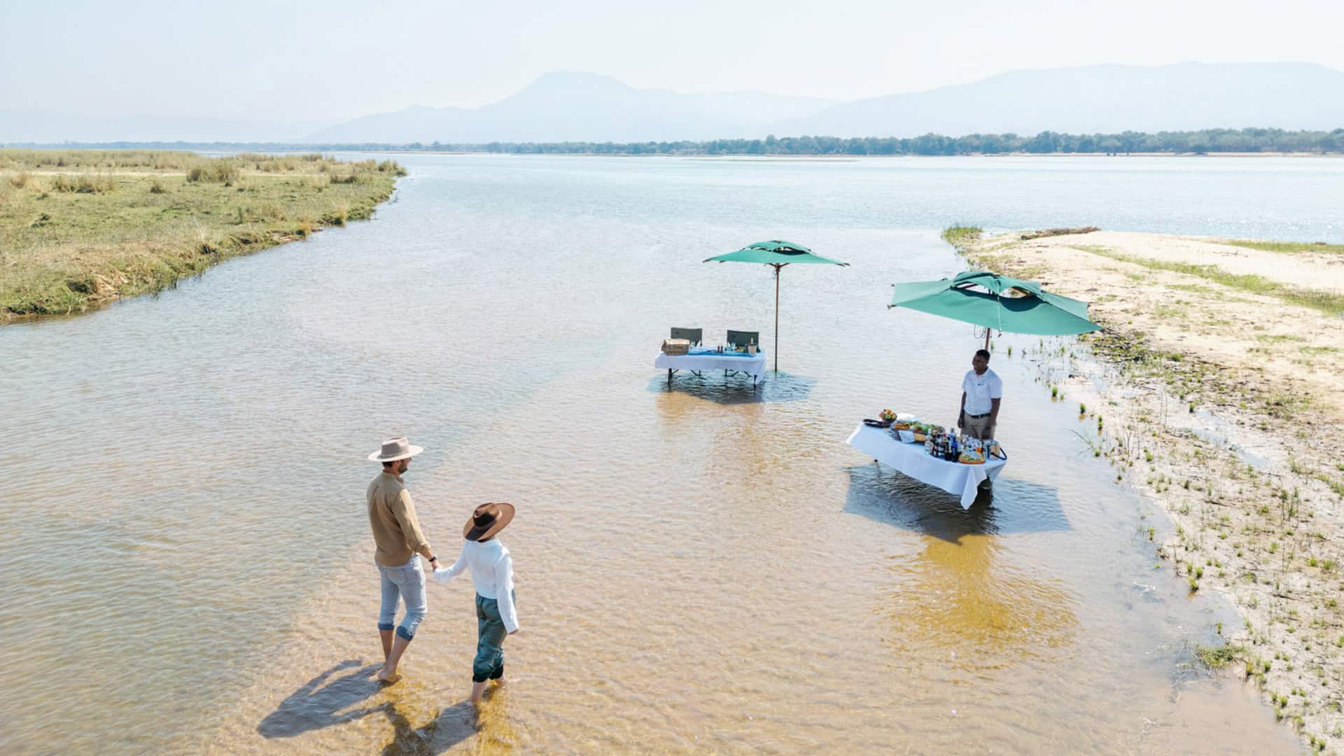 A couple holding hands and walking in the shallows of the river towards the picnic setup to be served with Time + Tide in Zambia